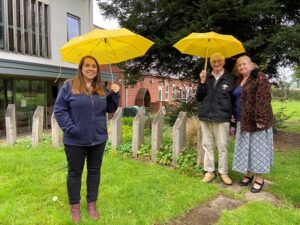 Lucy Perkins with Jim and Ruth Wood holding yellow umbrellas in the gardens of Primrose Hospice.