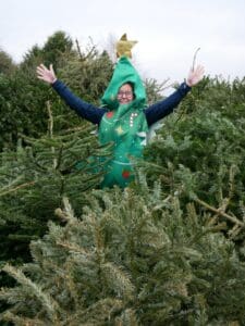 A person dressed as a christmas tree stood in the middle of a pile of real christmas trees.